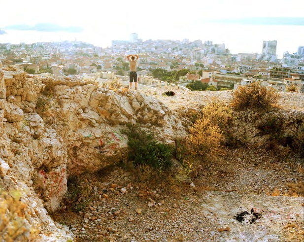 A figure standing on a rocky rise overlooking a dense coastal city