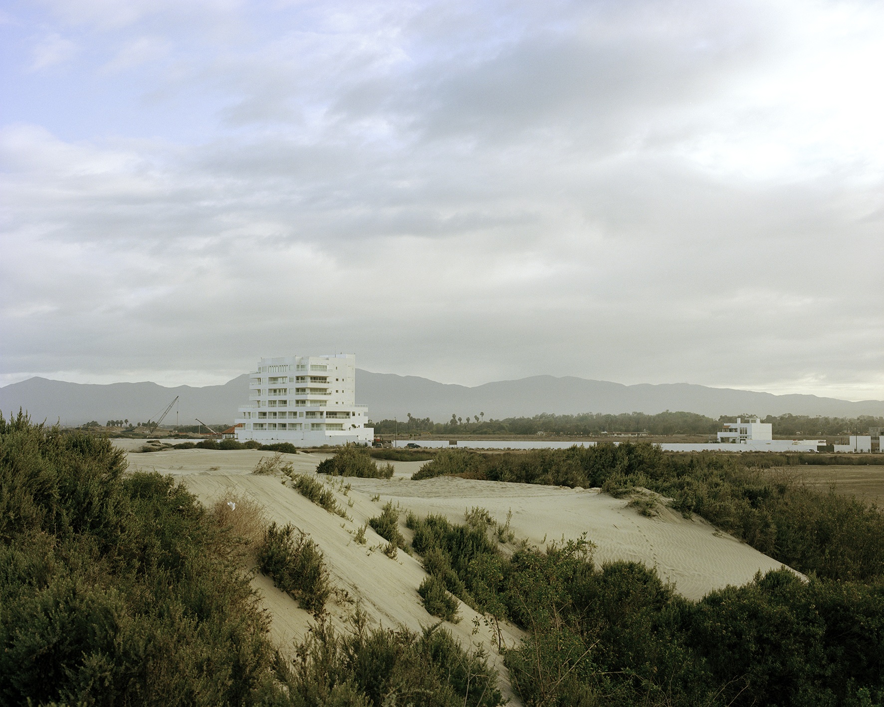 Wetland and sandy terrain surrounding an isolated apartment building beneath a pale sky